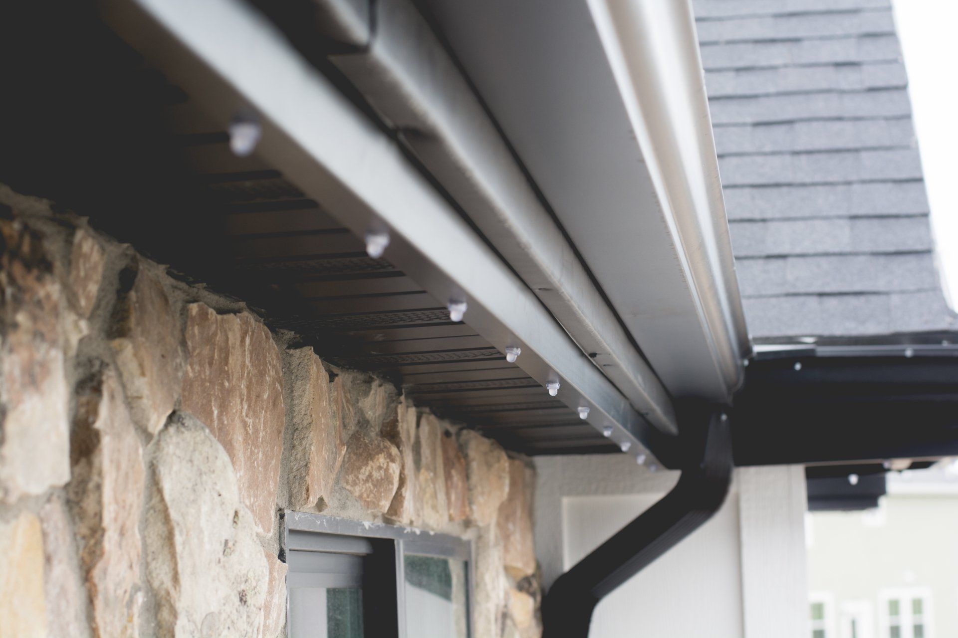 Close-up of roof eaves, gutter, and downspout beside a stone wall and window