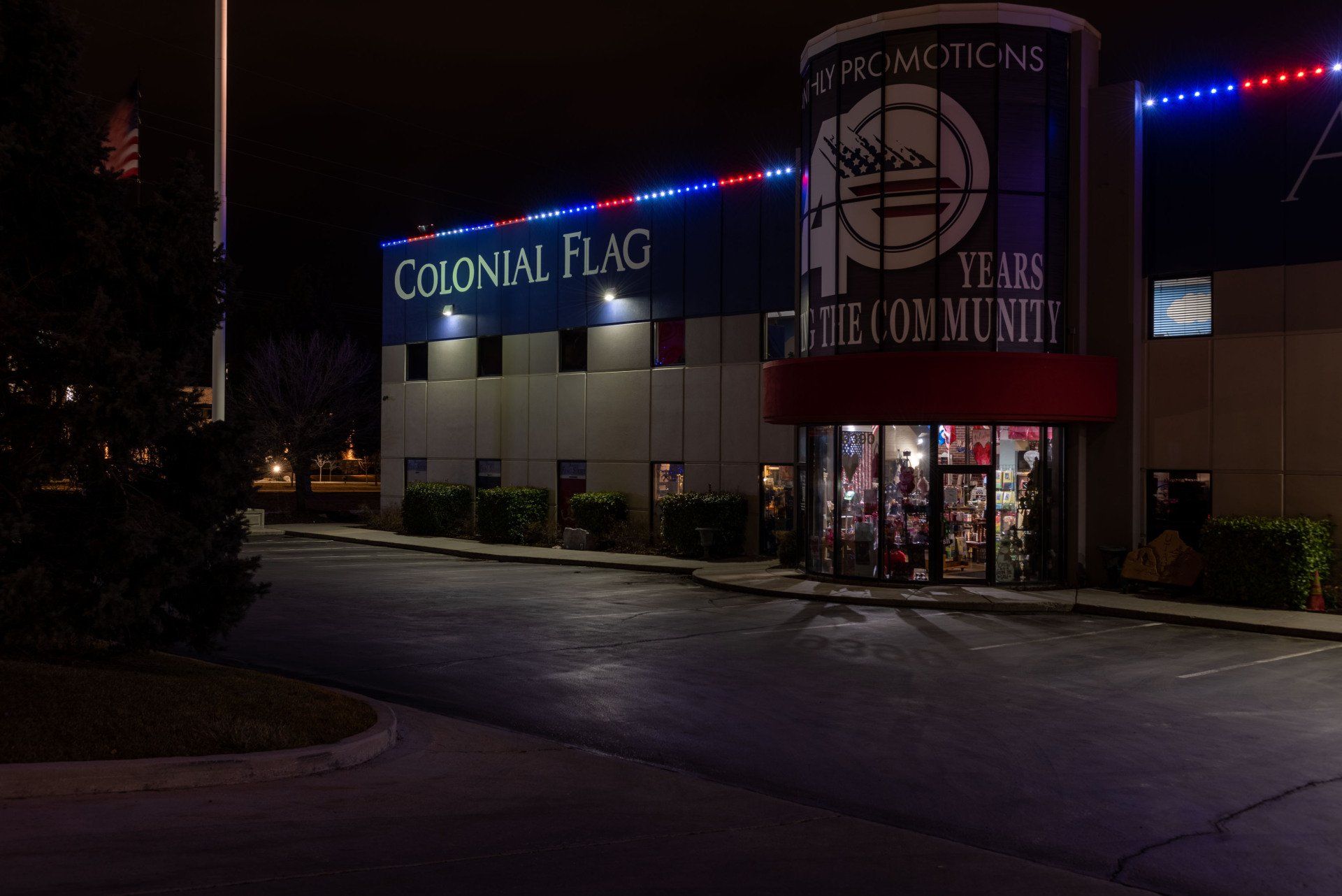 Nighttime exterior of Colonial Flag storefront with illuminated signs and a lit parking lot