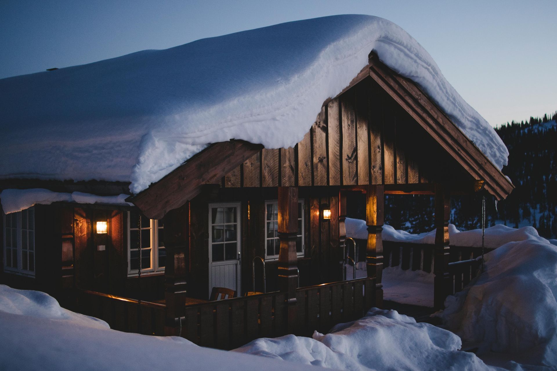 Snow-covered wooden cabin at dusk, lights glowing from windows and porch.