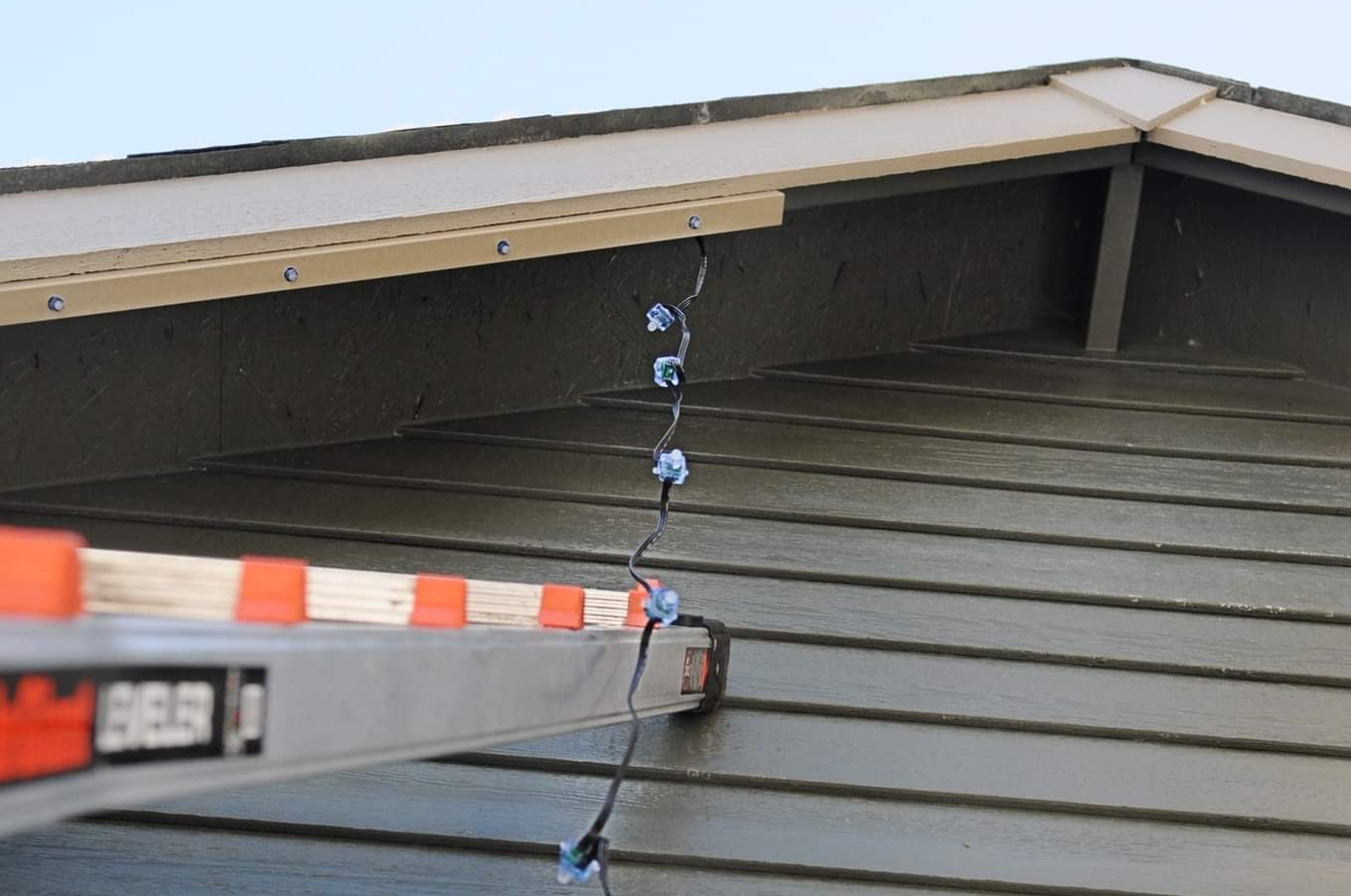 Ladder against a gray house roof with blue holiday lights hanging from the eaves