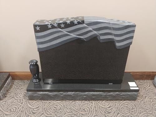 Black granite tombstone with a carved, draped American flag design, resting on a pedestal in an indoor showroom.