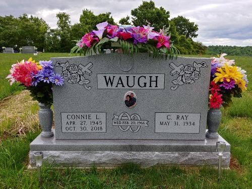 A grey granite Waugh family headstone in a cemetery, featuring floral arrangements and dates for Connie and C. Ray.