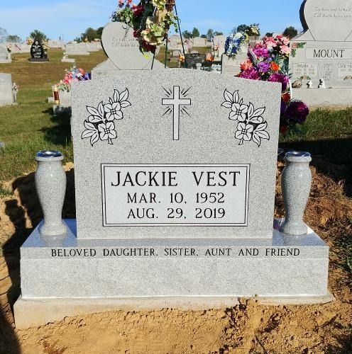A gray granite headstone for Jackie Vest (1952–2019) with floral carvings and a cross in a cemetery setting.