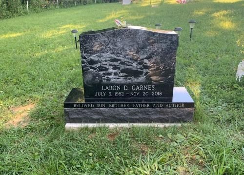 A black granite gravestone engraved with a mountain stream scene, name Lakon D. Garnes, and dates in a grassy cemetery.