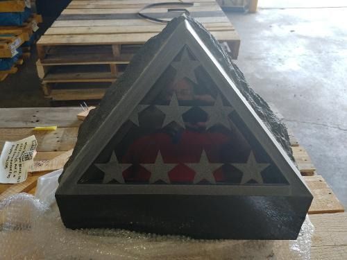 A triangular, dark stone memorial with an inset glass panel displaying American flag stars, resting on a wooden pallet.