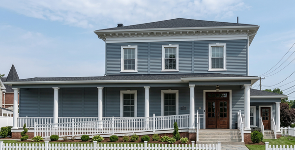 A blue, two-story historic house with a full-width covered porch, white columns, and a wooden wheelchair ramp.