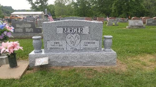A gray granite tombstone with the name BERGER engraved, centered in a grassy cemetery with flowers nearby.