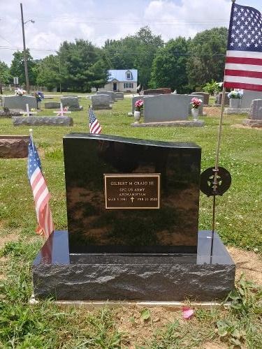 A polished black headstone with a brass plaque in a cemetery, flanked by two small American flags on a sunny day.