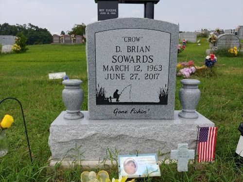 A gray granite tombstone for D. Brian Sowards in a cemetery, featuring an etched fishing scene and the words 