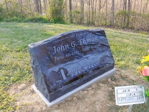 A dark grey granite headstone engraved with a landscape scene, names, and dates, set in a grassy cemetery.