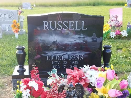 A black granite headstone with the name RUSSELL and dates Nov 22, 1939 – Feb 17, 2019, surrounded by colorful flowers.