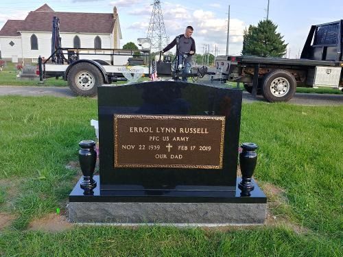 A person installing a black granite headstone for Errol Lynn Russell in a cemetery with a church in the background.
