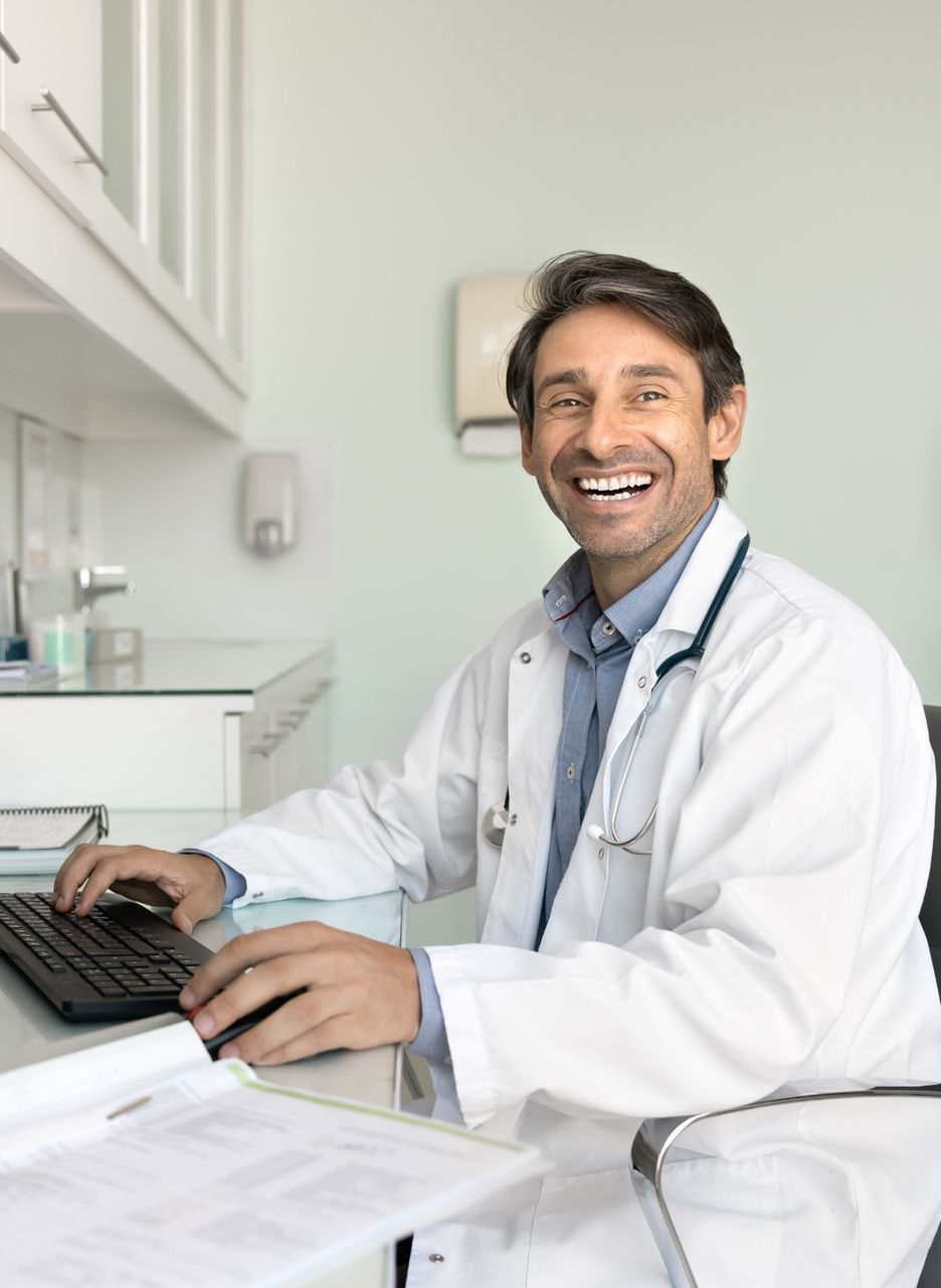 Doctor in a white coat smiling at a desk with a laptop in a bright clinic office