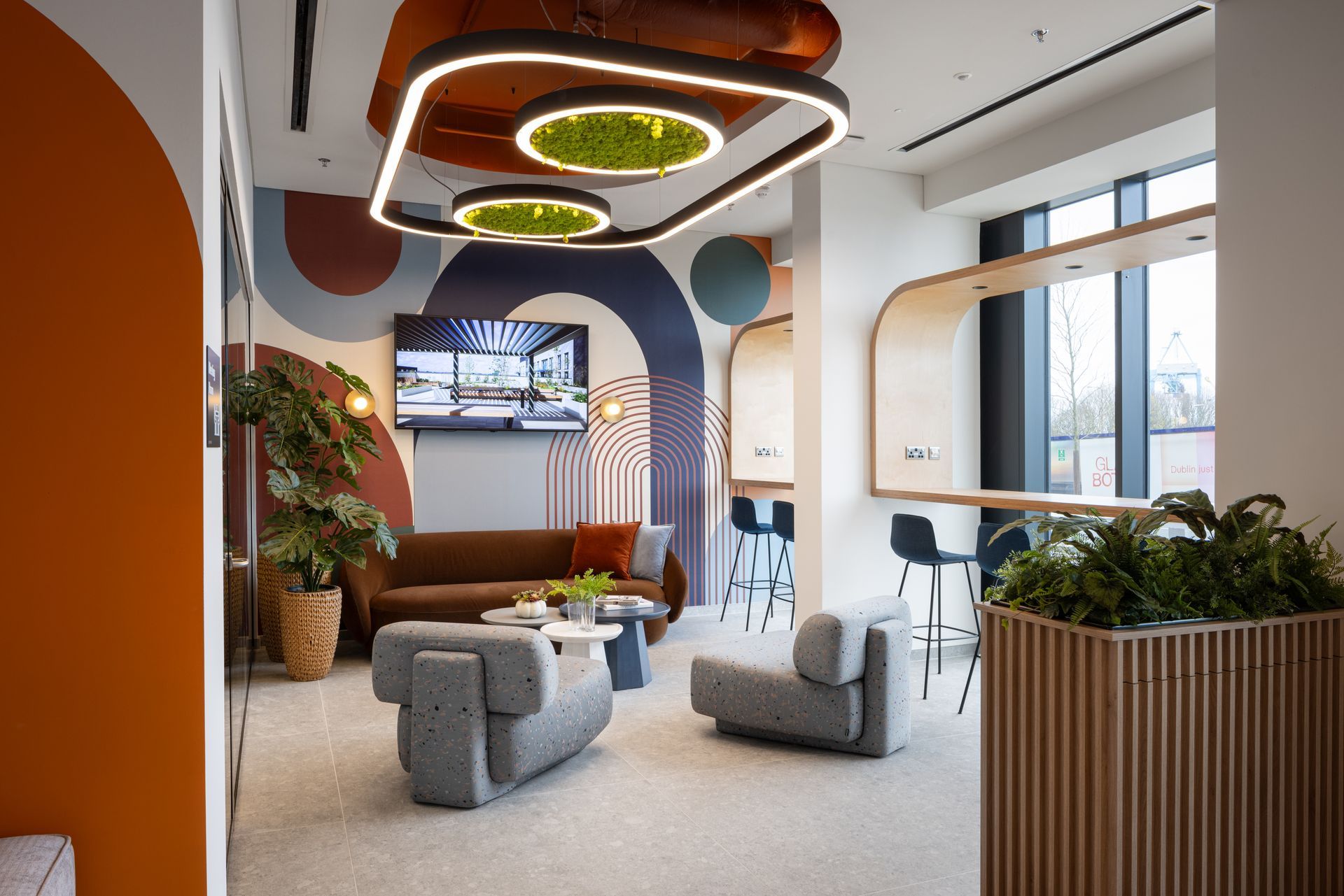 Modern lounge with curved ceiling lights, gray seating, plants, and large windows in a bright office space
