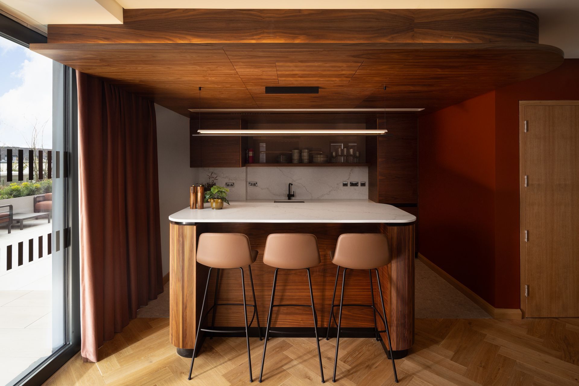 Modern wood-paneled kitchenette with three stools, white counter, and large window light