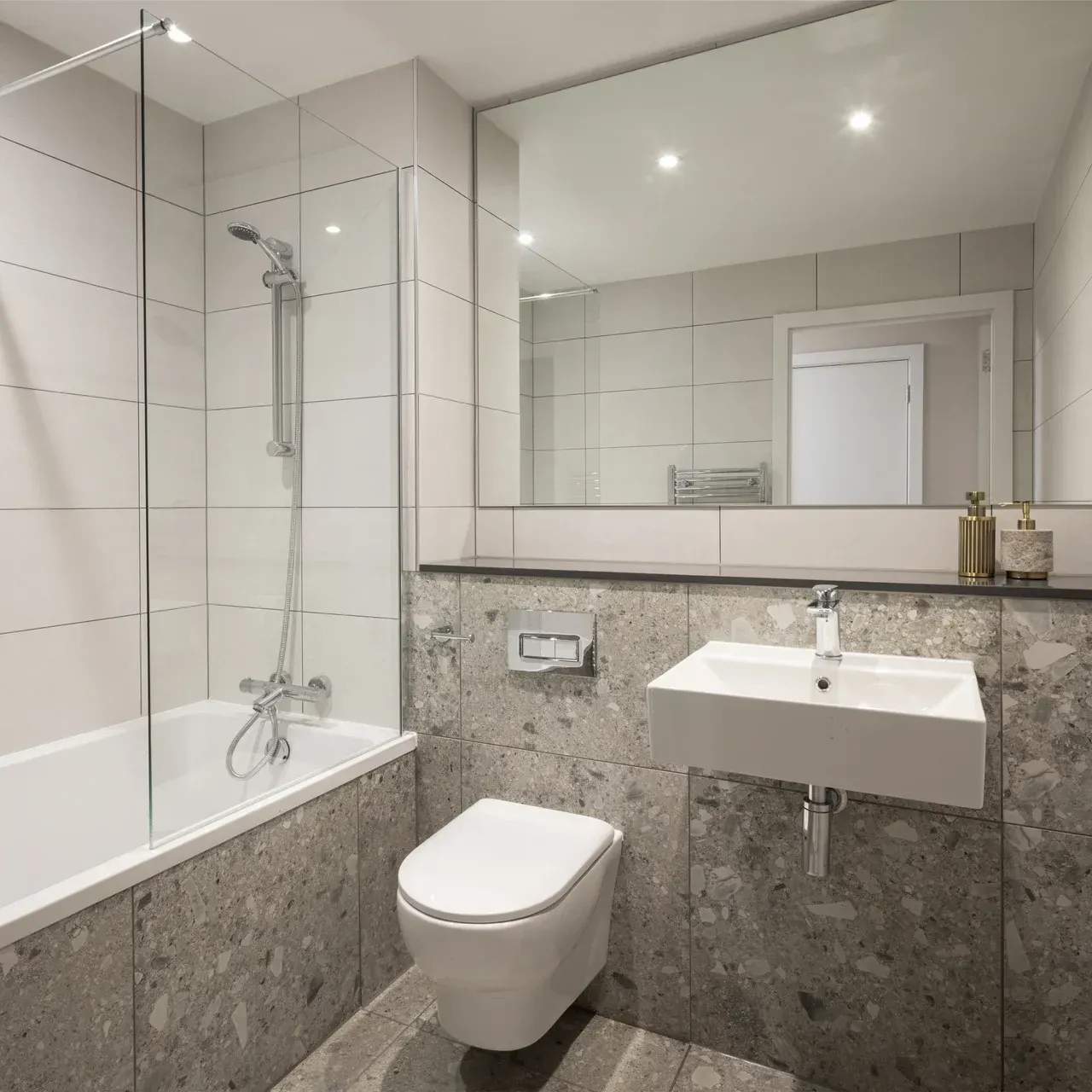Compact modern bathroom with white fixtures, gray stone tiles, bathtub, wall-mounted sink, and large mirror.