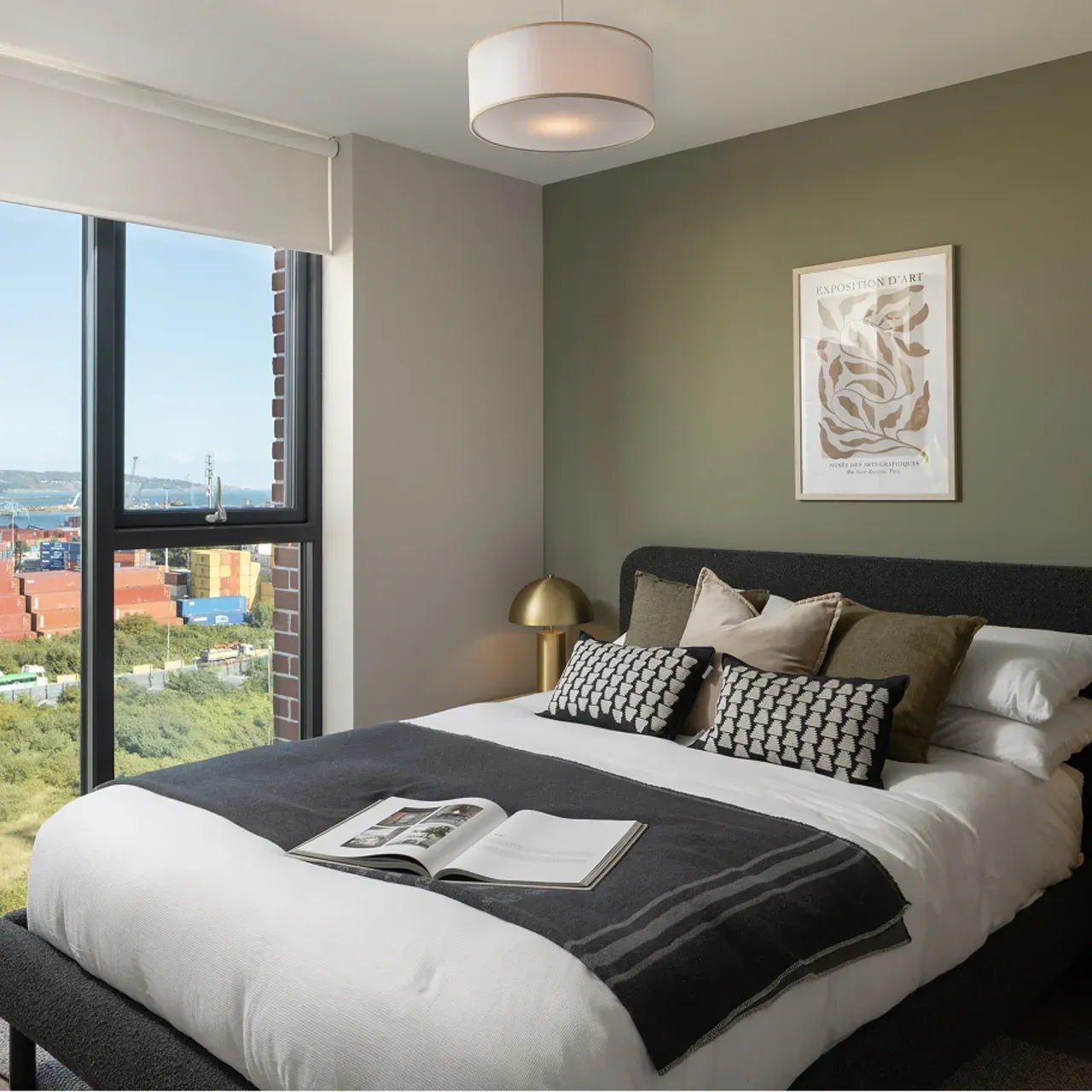 Modern bedroom with a neatly made bed, desk, and city view through a large window