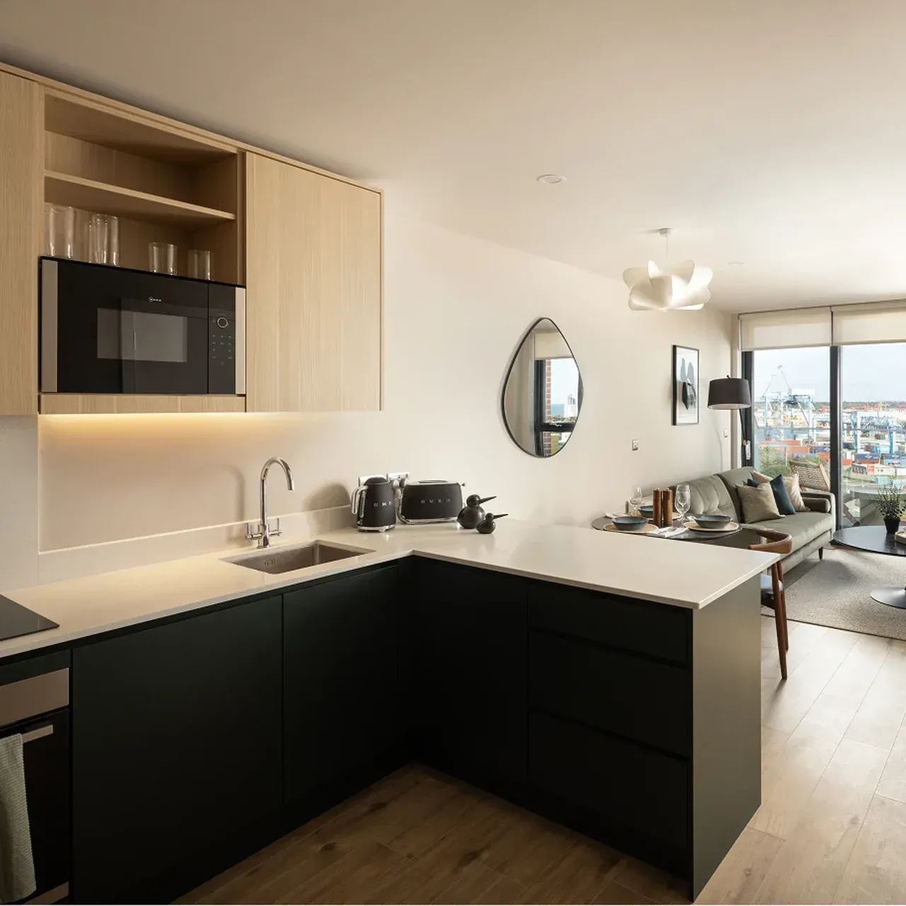 Modern kitchen with black cabinets, light wood shelving, and a view into a bright living room.