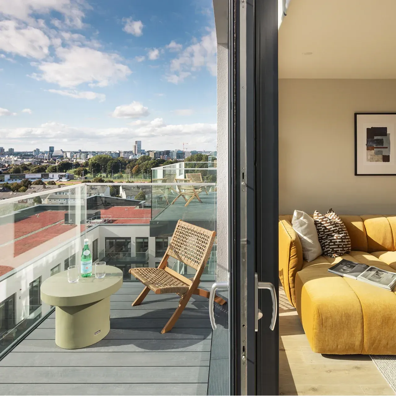 Balcony with chairs and table beside a sunlit living room, overlooking a city skyline.