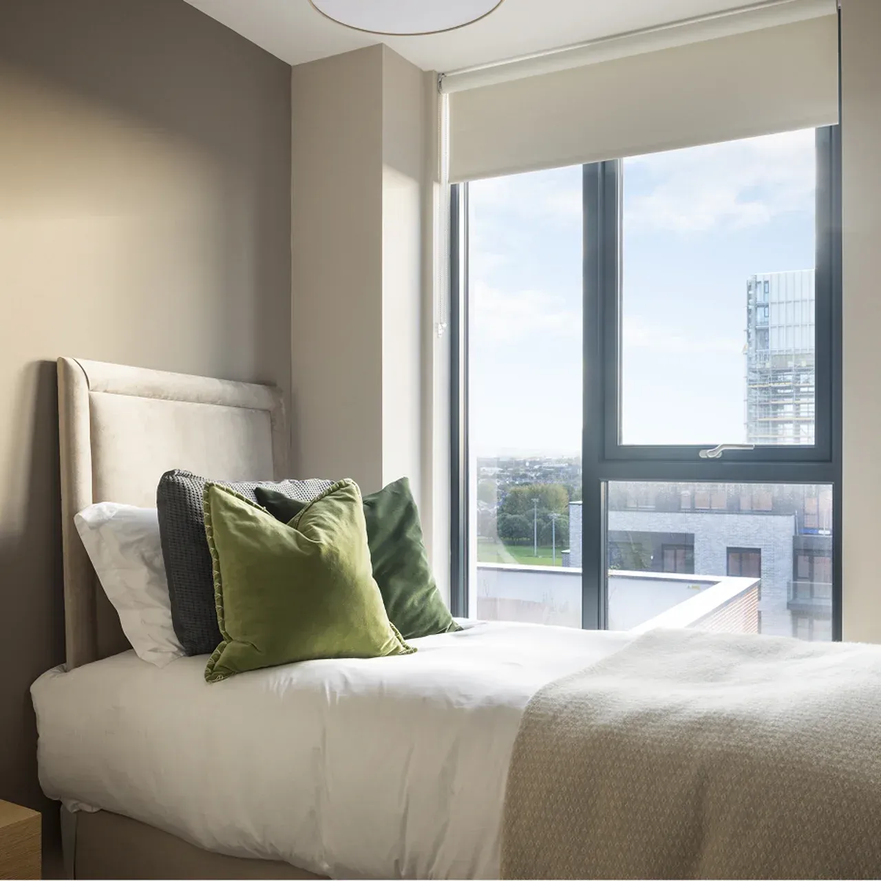 Neatly made bed beside a large window in a bright modern bedroom with green accent pillows.
