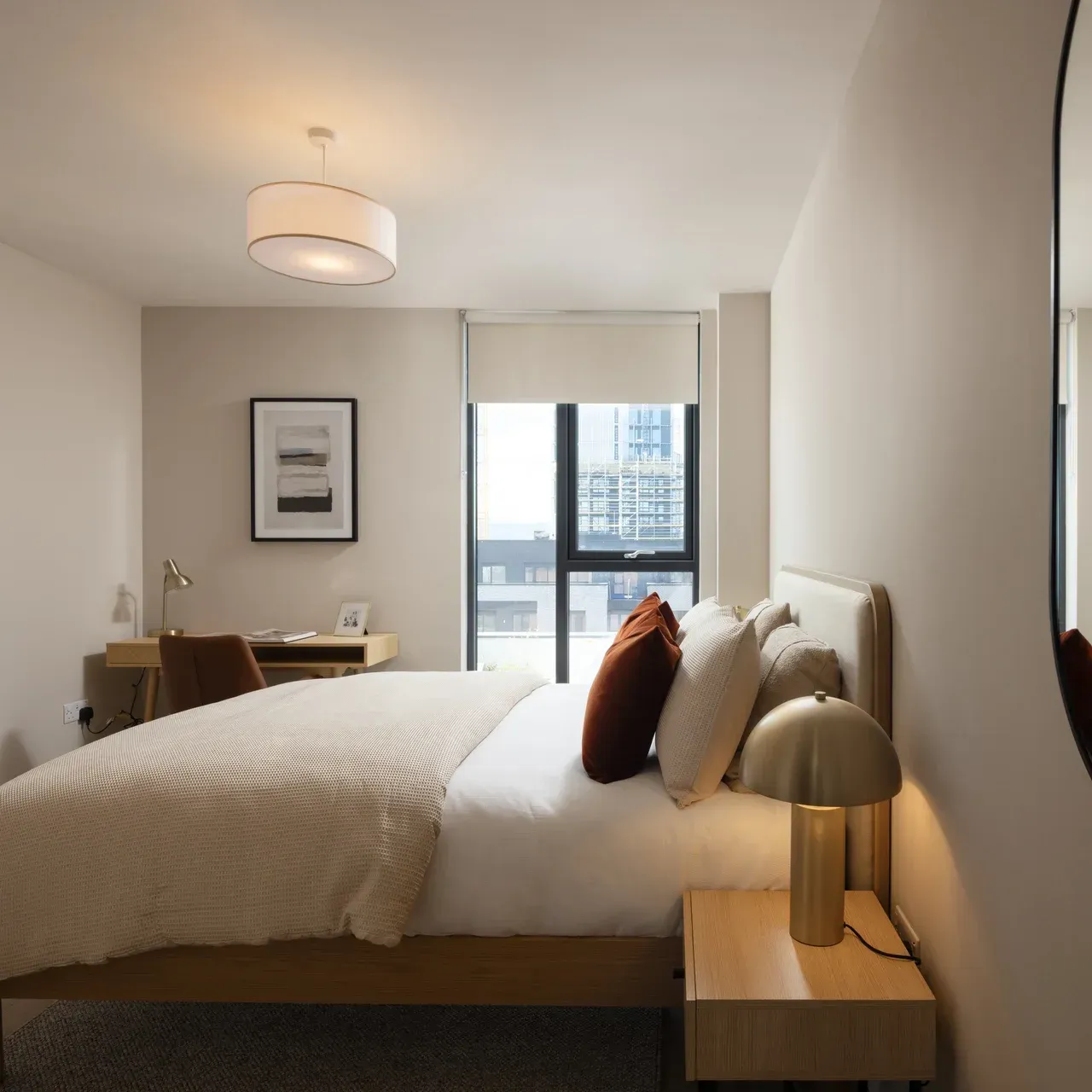 Modern bedroom with a neatly made bed, desk, and city view through a large window