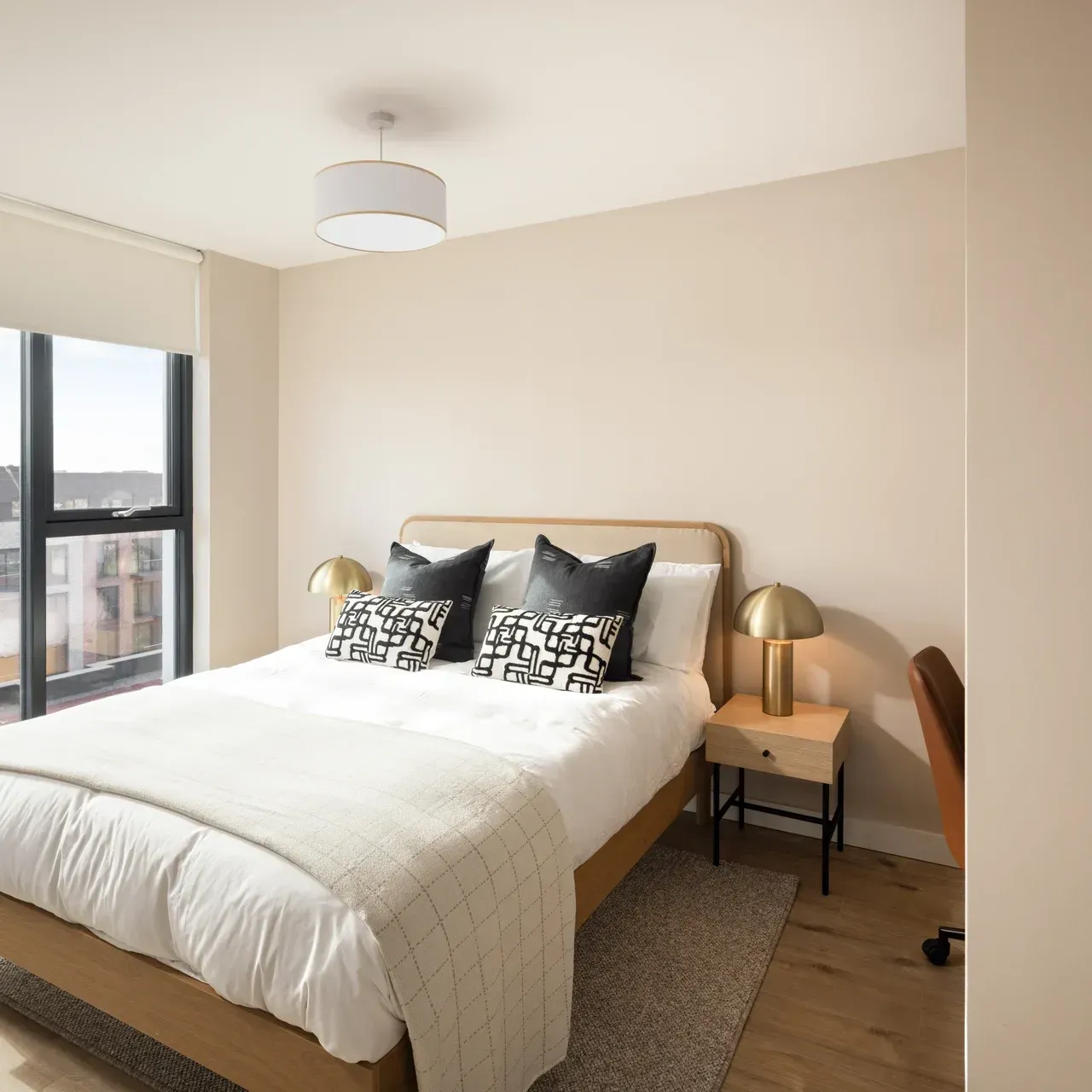 Bright bedroom with a made bed, bedside lamps, window light, and beige walls.