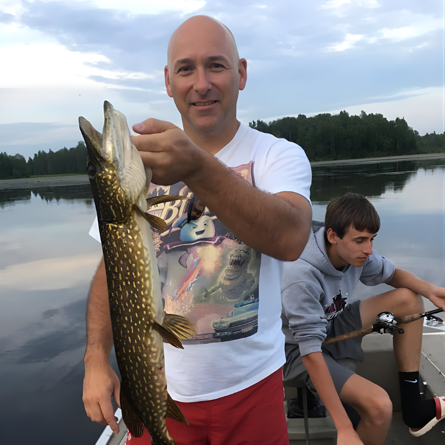 Man holding a large fish by a calm lakeshore, with another person sitting in a boat behind him.