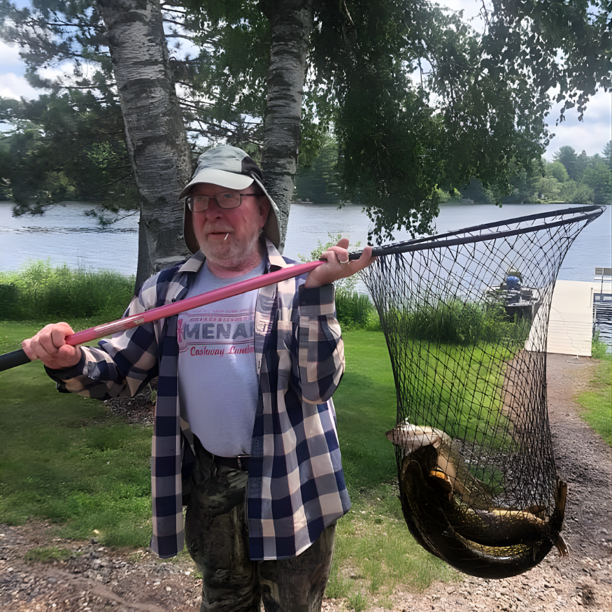 Person holding a fishing net with a large fish beside a lakeside dock, standing under trees.