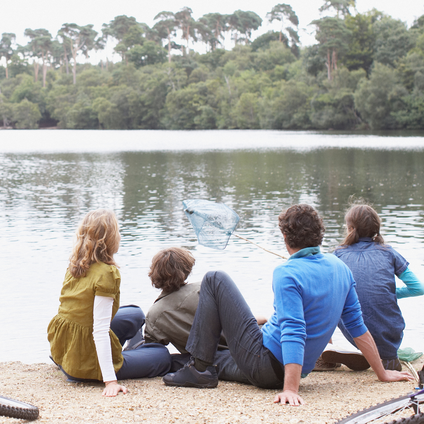 Five people sit by a lake, looking at a splash in the water with trees across the shore.