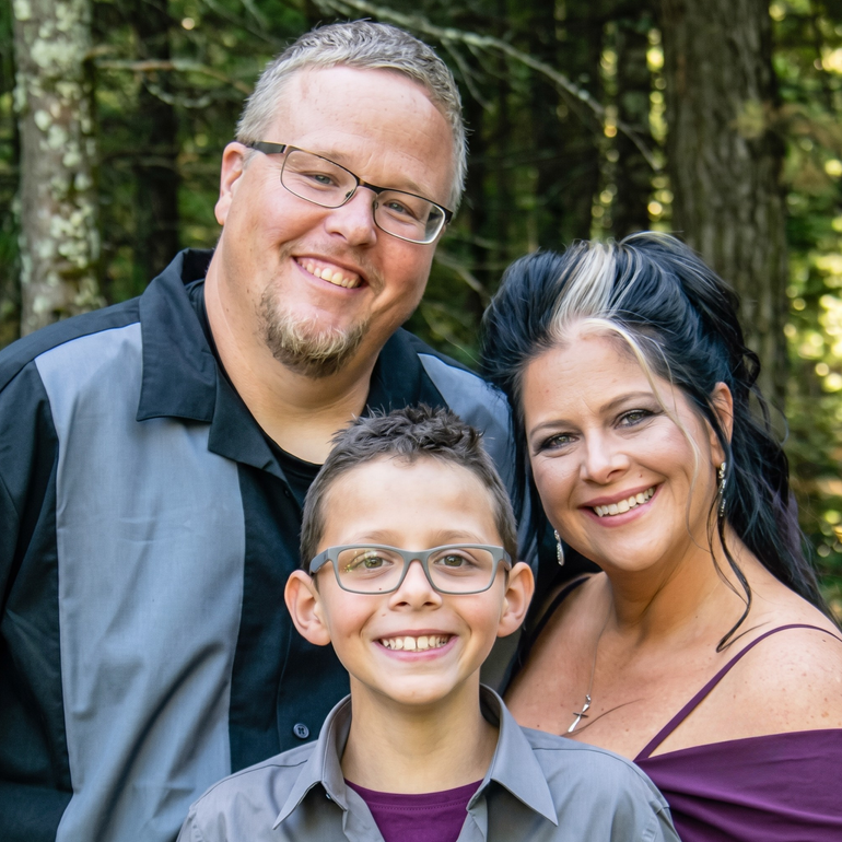 Smiling family of three posing outdoors in a wooded area.