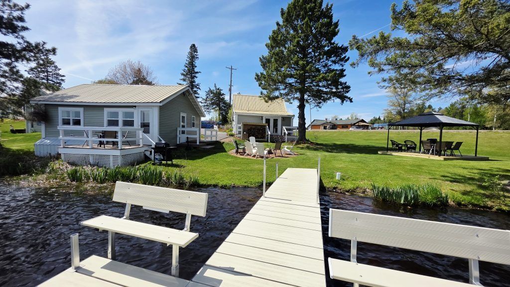 Lakeside cabin with dock, benches, picnic shelter, and trees under a clear blue sky