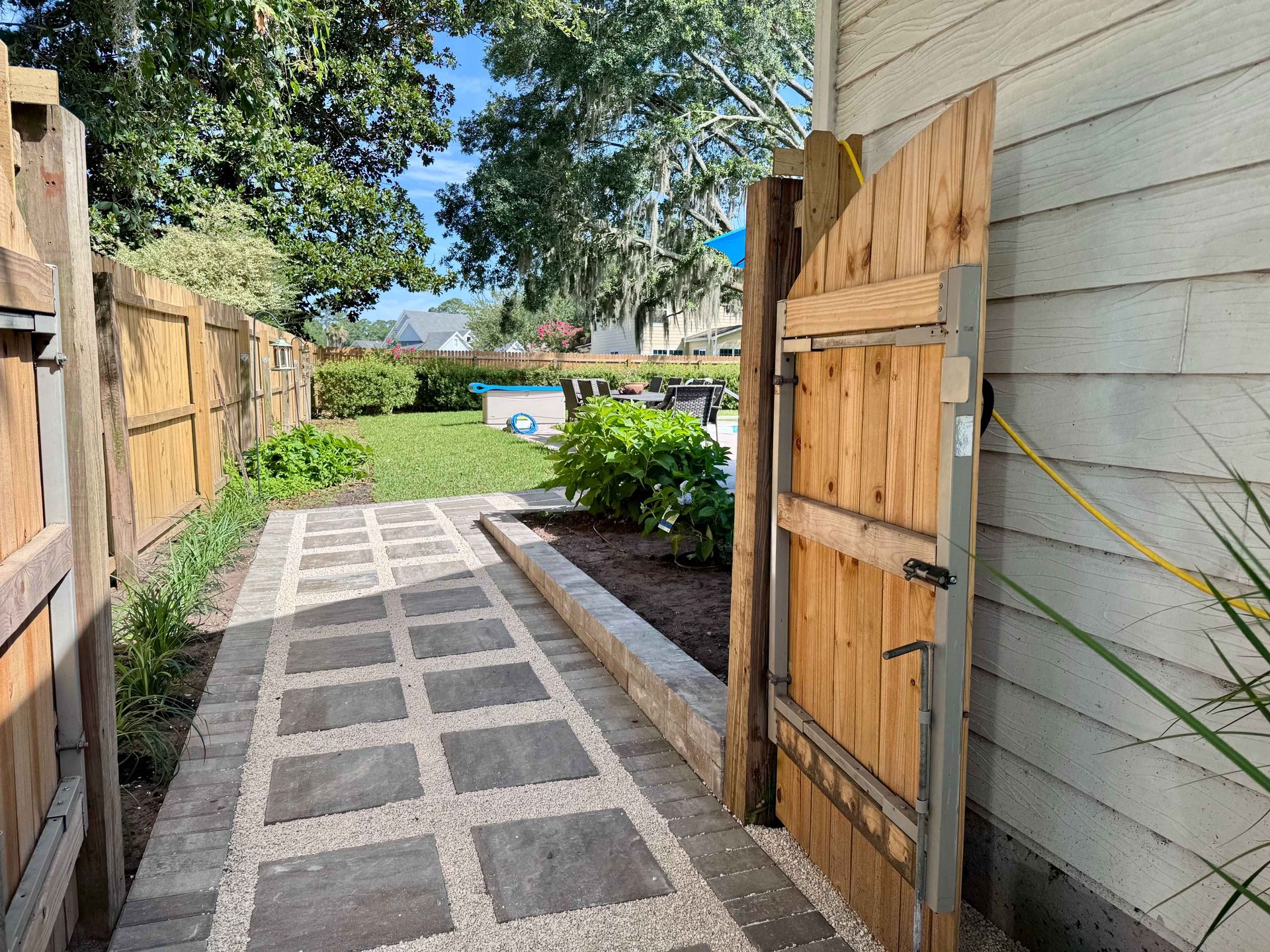 Wooden gate open to a paved walkway, leading to a grassy yard with a pool, bordered by a wooden fence.