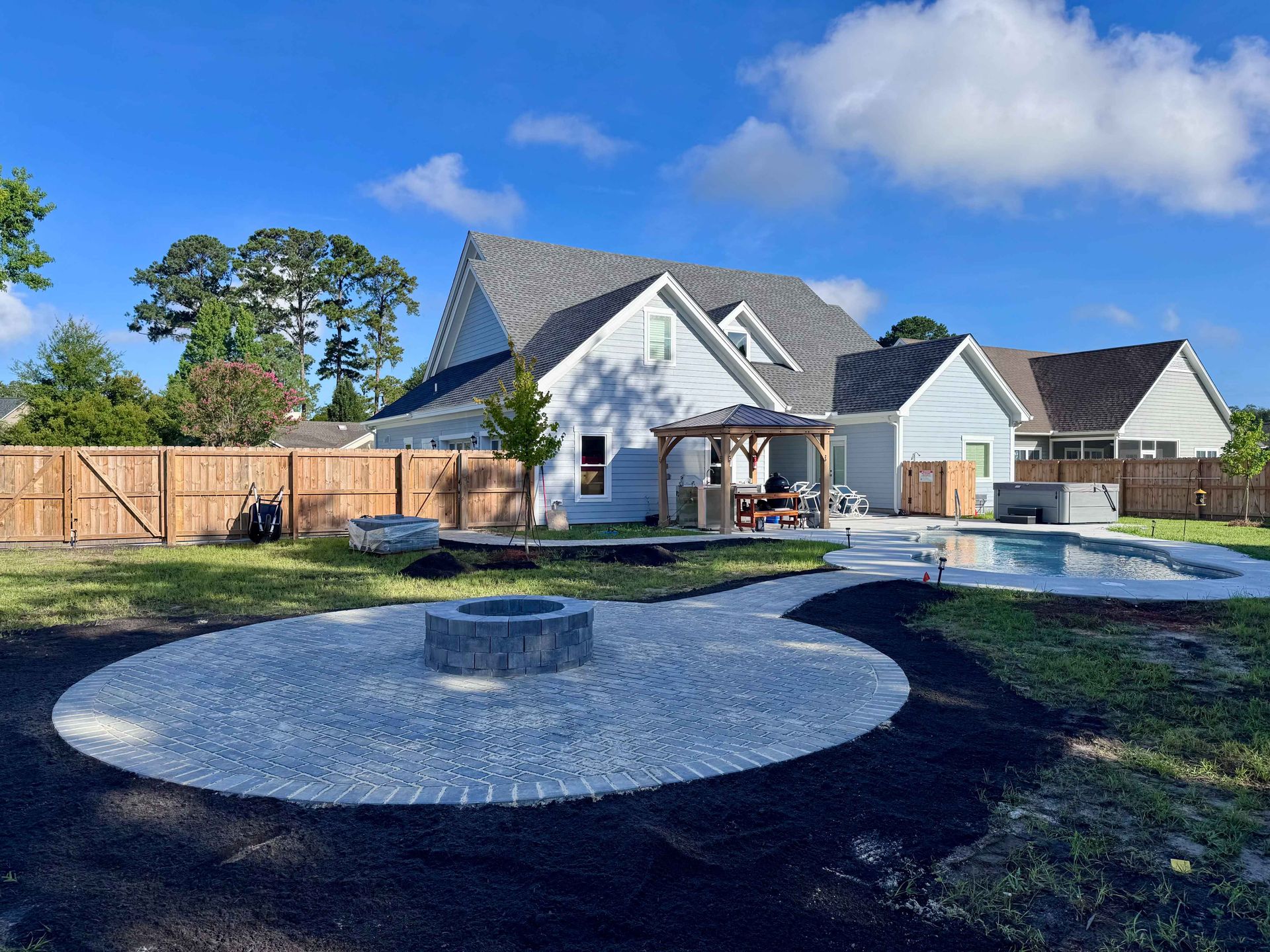 Backyard with a pool, fire pit, patio, and light blue house with wooden fence and blue sky.