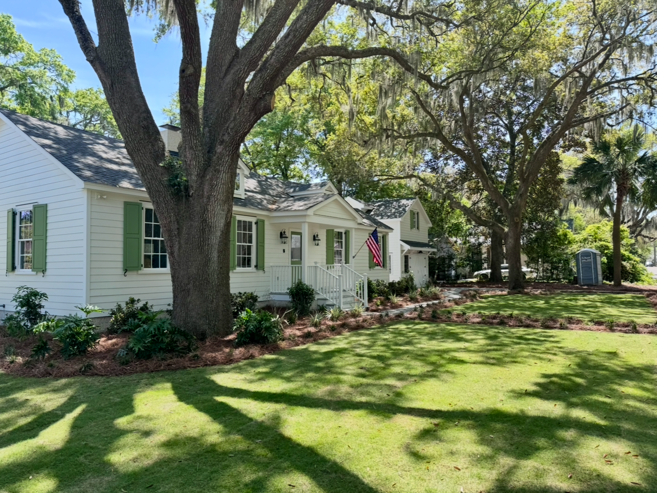 A white house with green shutters and a large tree in front of it.