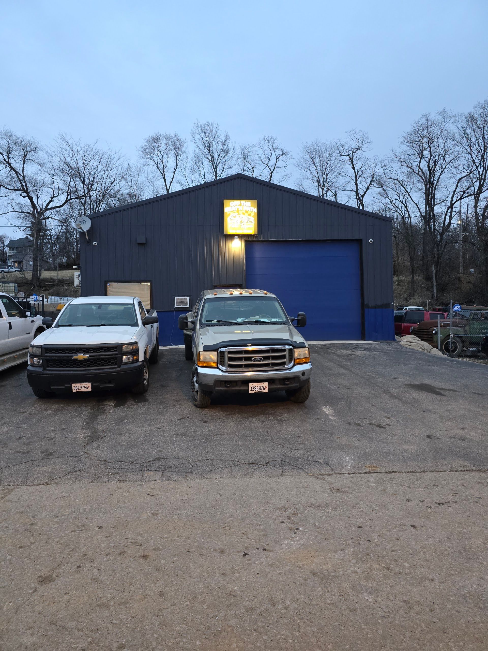 Two pickup trucks parked in front of a blue garage at dusk | Off The Beaten Path Automotive LLC