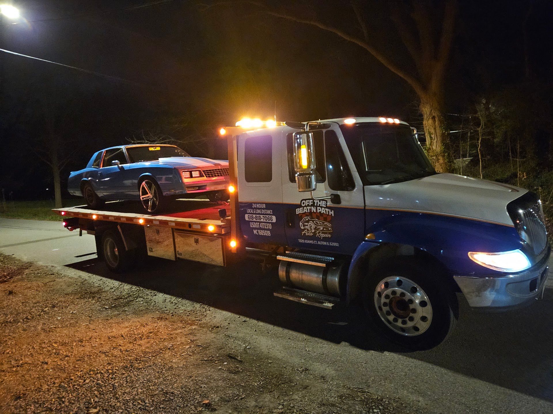 Tow truck hauling a dark car at night, with flashing amber lights on a roadside | Off The Beaten Path Automotive LLC