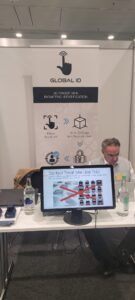 Stand d'exposition avec une bannière « Global ID » et un homme derrière une table présentant un écran avec des visuels de produits.