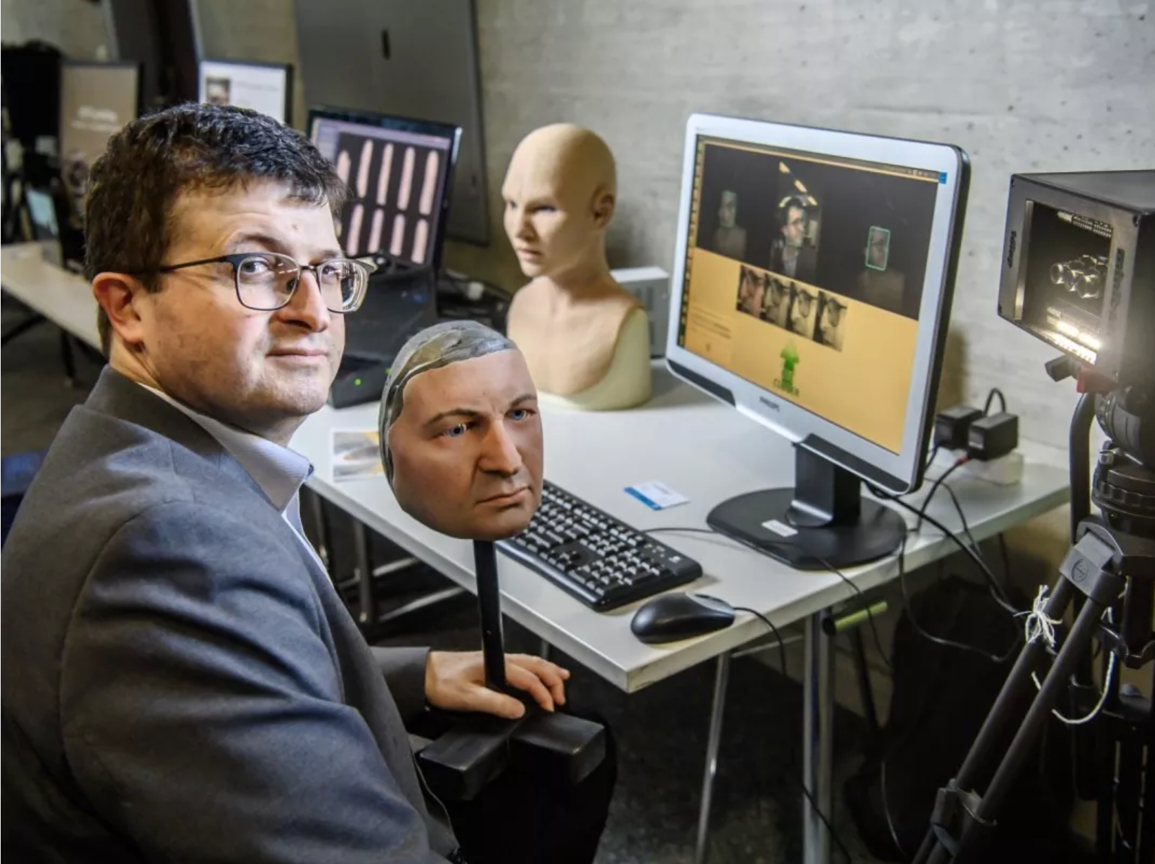 Un homme est assis à un bureau d'ordinateur avec deux visages découpés, une tête de mannequin et un écran affichant une page web.