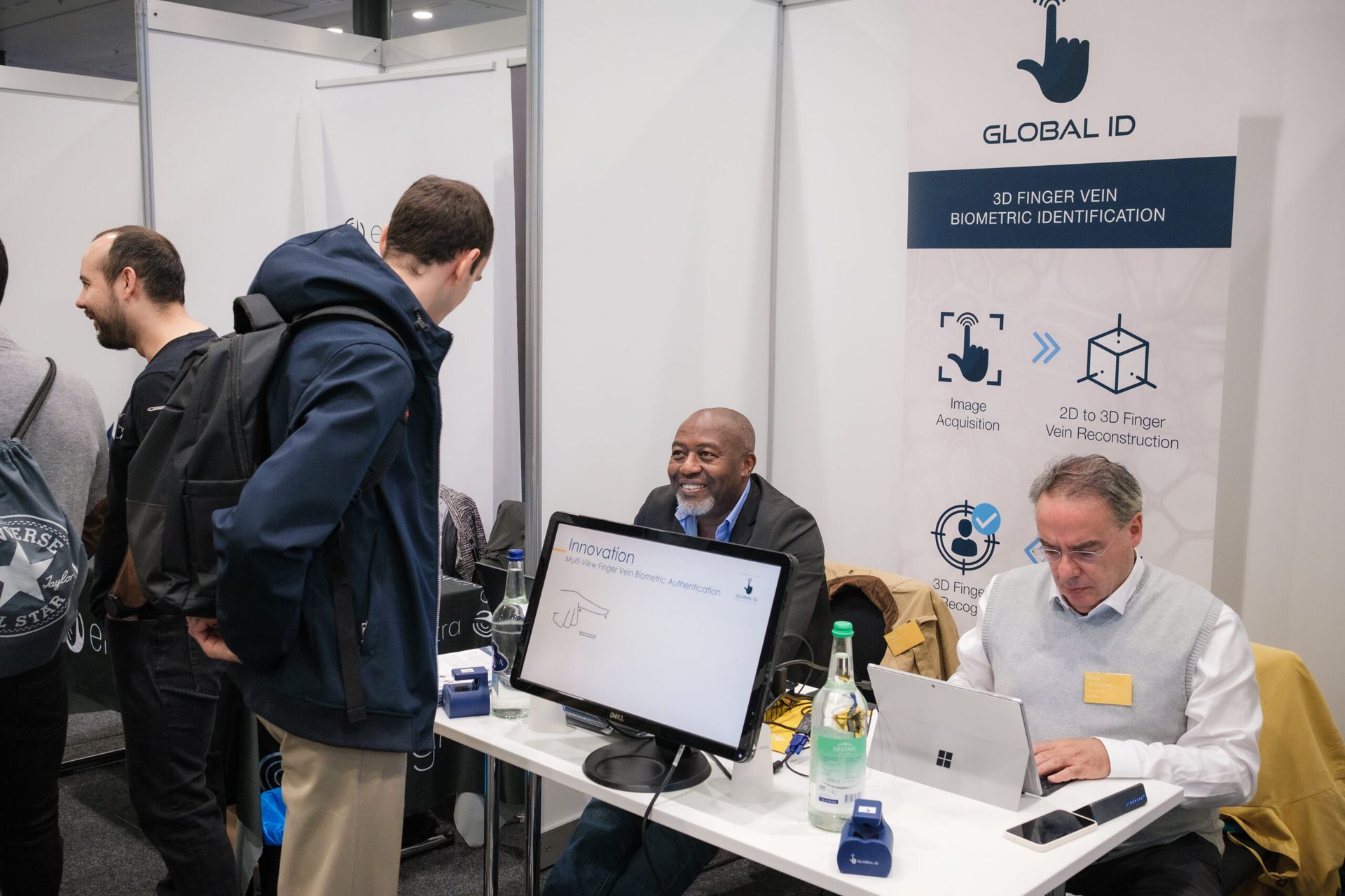 Des personnes sur un stand lors d'un salon professionnel, avec deux hommes assis à une table utilisant des ordinateurs portables et un écran à proximité.