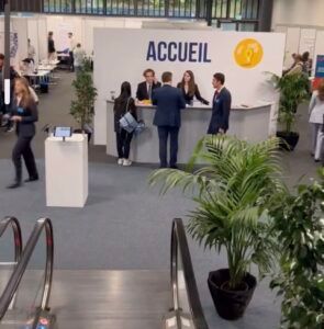 Zone d'accueil avec des personnes debout à un comptoir d'enregistrement sous une grande enseigne « ACCUEIL », plantes au premier plan.