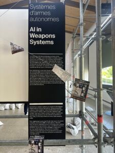 Panneau d'exposition du musée sur l'IA dans les systèmes d'armement, à côté d'échafaudages métalliques et de panneaux suspendus.