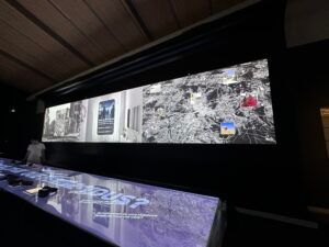 Salle d'exposition obscure avec un grand écran panoramique diffusant des cartes et des images de la ville, à côté d'un écran lumineux au sol.