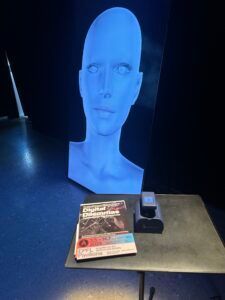 Buste bleu imprimé en 3D posé sur une table à côté d'un magazine et d'un petit appareil dans une pièce sombre