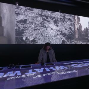Une personne dans une pièce faiblement éclairée interagit avec une longue table éclairée sous un écran mural en noir et blanc