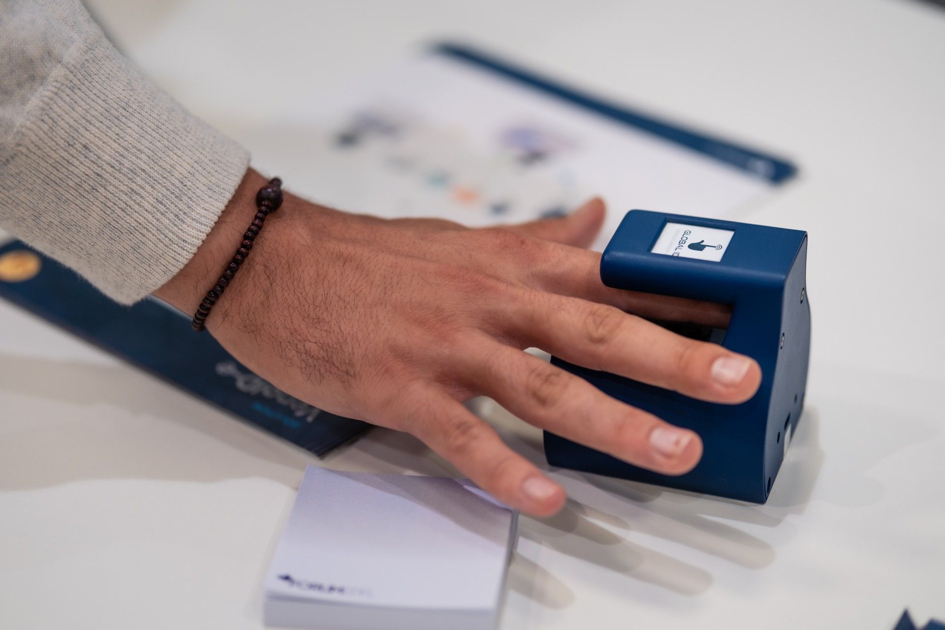 Une main appuie sur un appareil électronique bleu posé sur une table blanche, à côté d'un téléphone et de papiers.