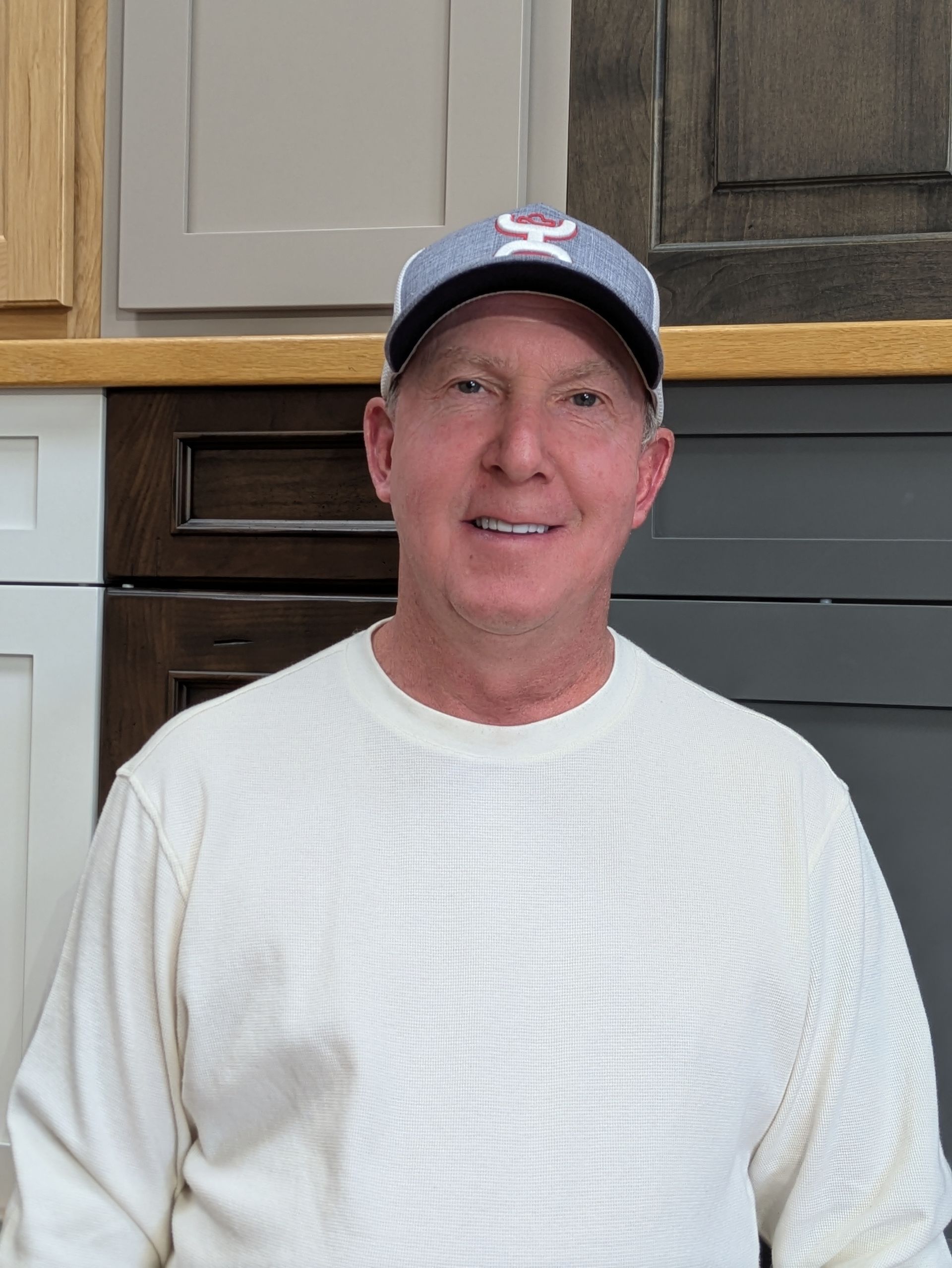 Man wearing a baseball cap and sweater, smiling, standing in front of cabinets.