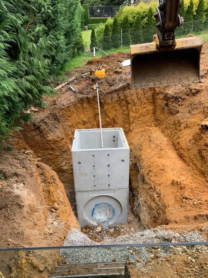 Excavator lowering concrete utility chamber into a dirt trench beside a green hedge