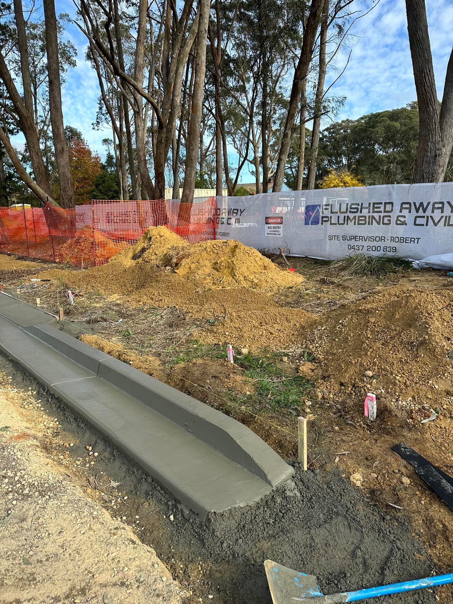 Construction site with orange fencing, dirt mound, curb, and trees beside a roadside.