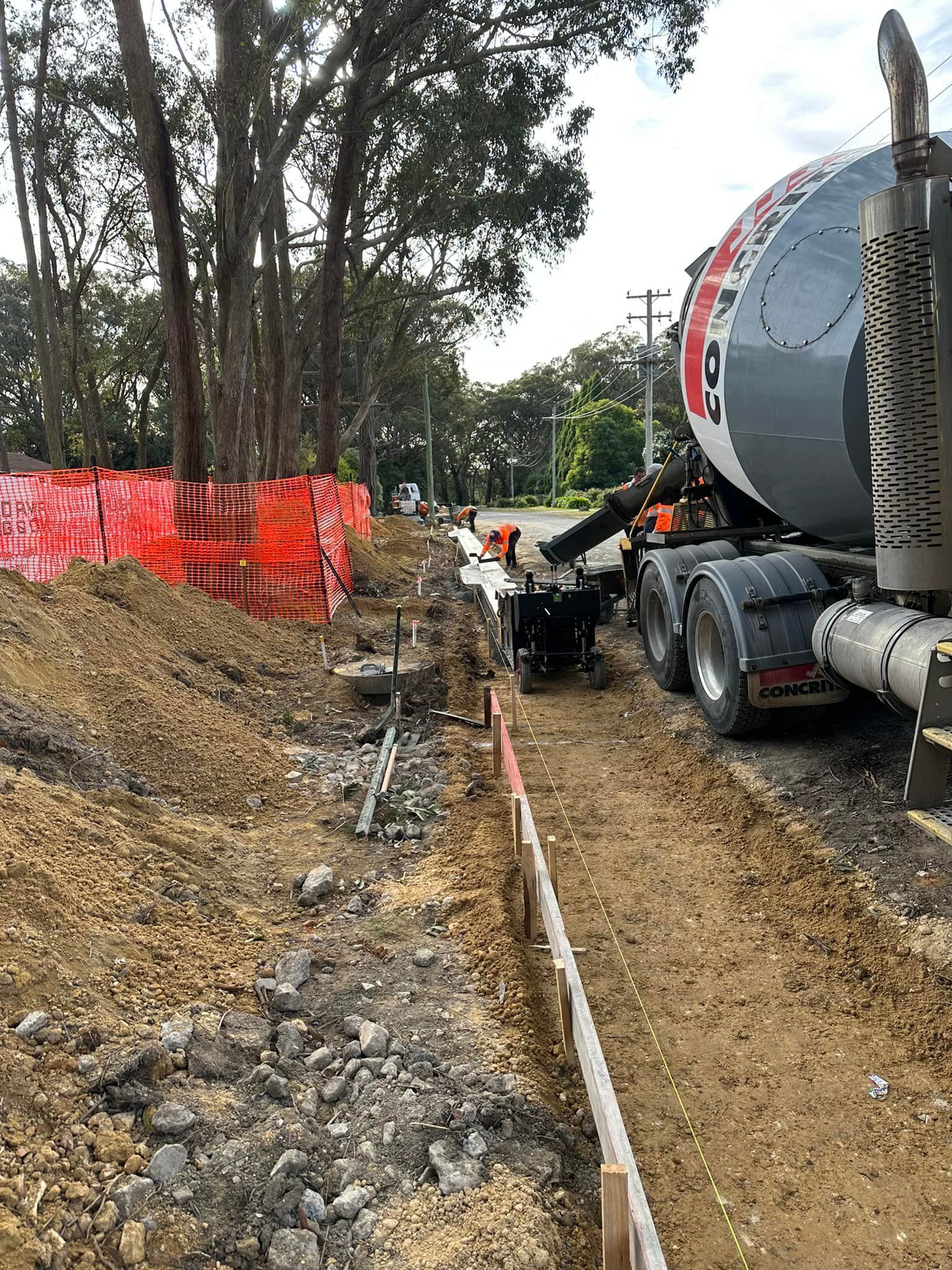 Concrete mixer truck beside trench with orange safety fencing and trees at a roadside worksite