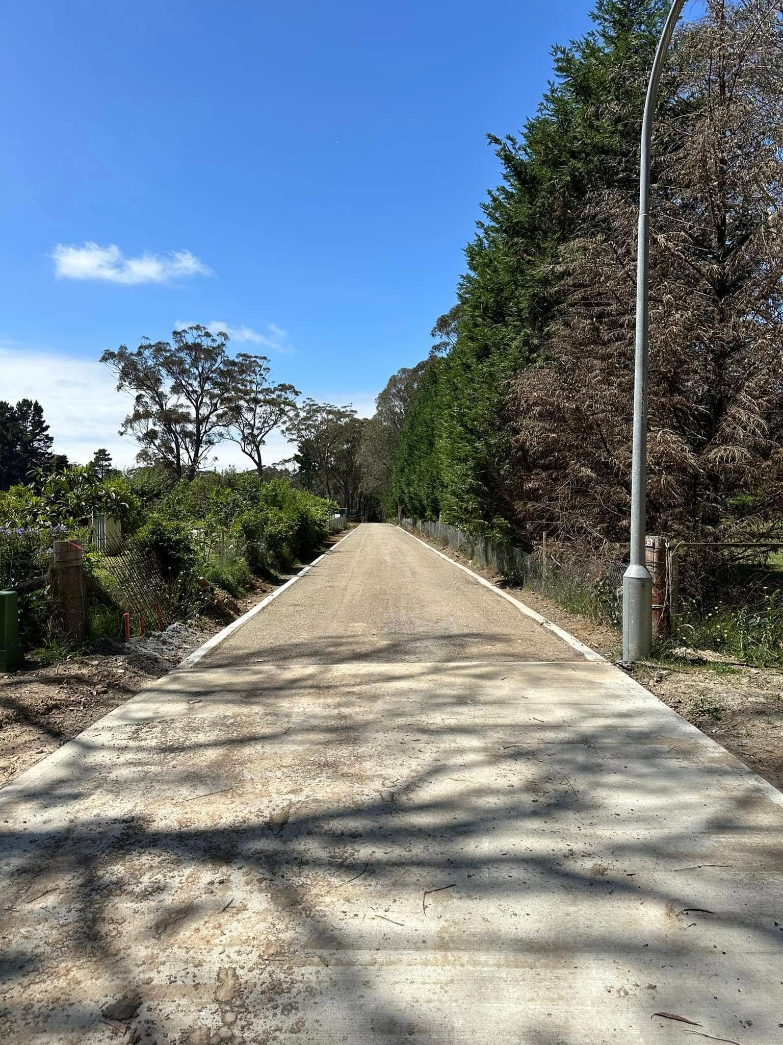Concrete path through a wooded area under a blue sky with a streetlight on the right